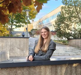 Elina yliopiston keskusaukiolle johtavien portaiden tasanteella, päärakennus ja puita taustalla.