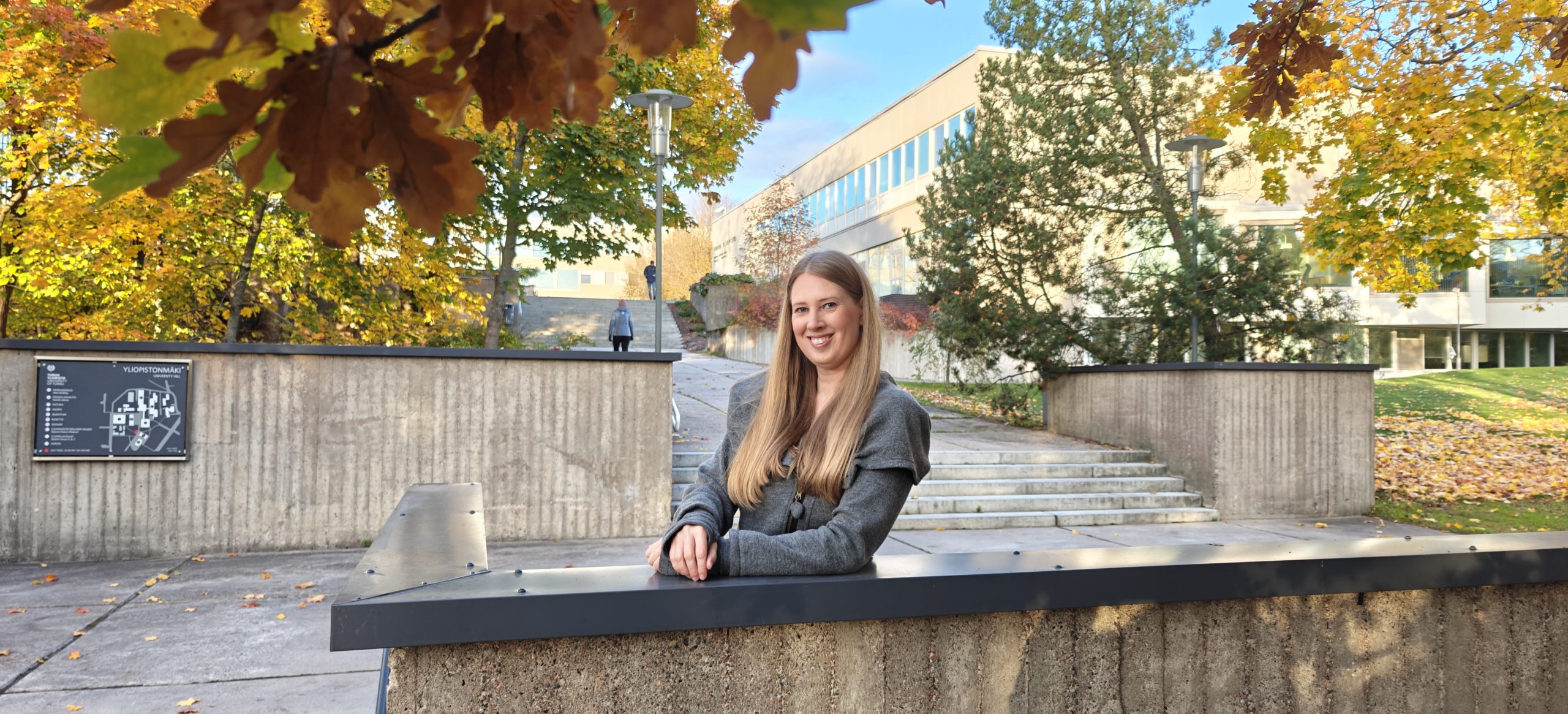 Elina yliopiston keskusaukiolle johtavien portaiden tasanteella, päärakennus ja puita taustalla.