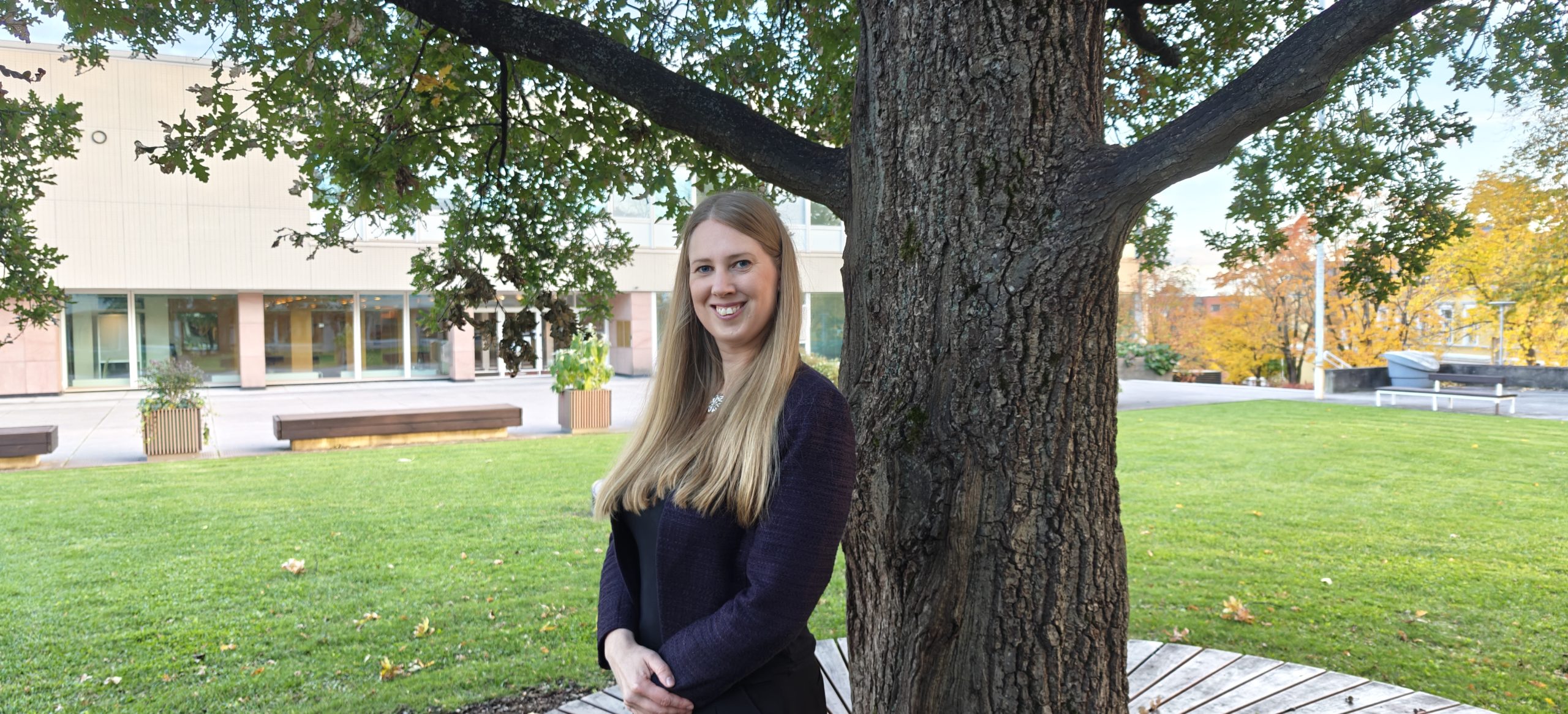 Elina yliopiston keskusaukiolla puun vieressä seisomassa, päärakennus taustalla