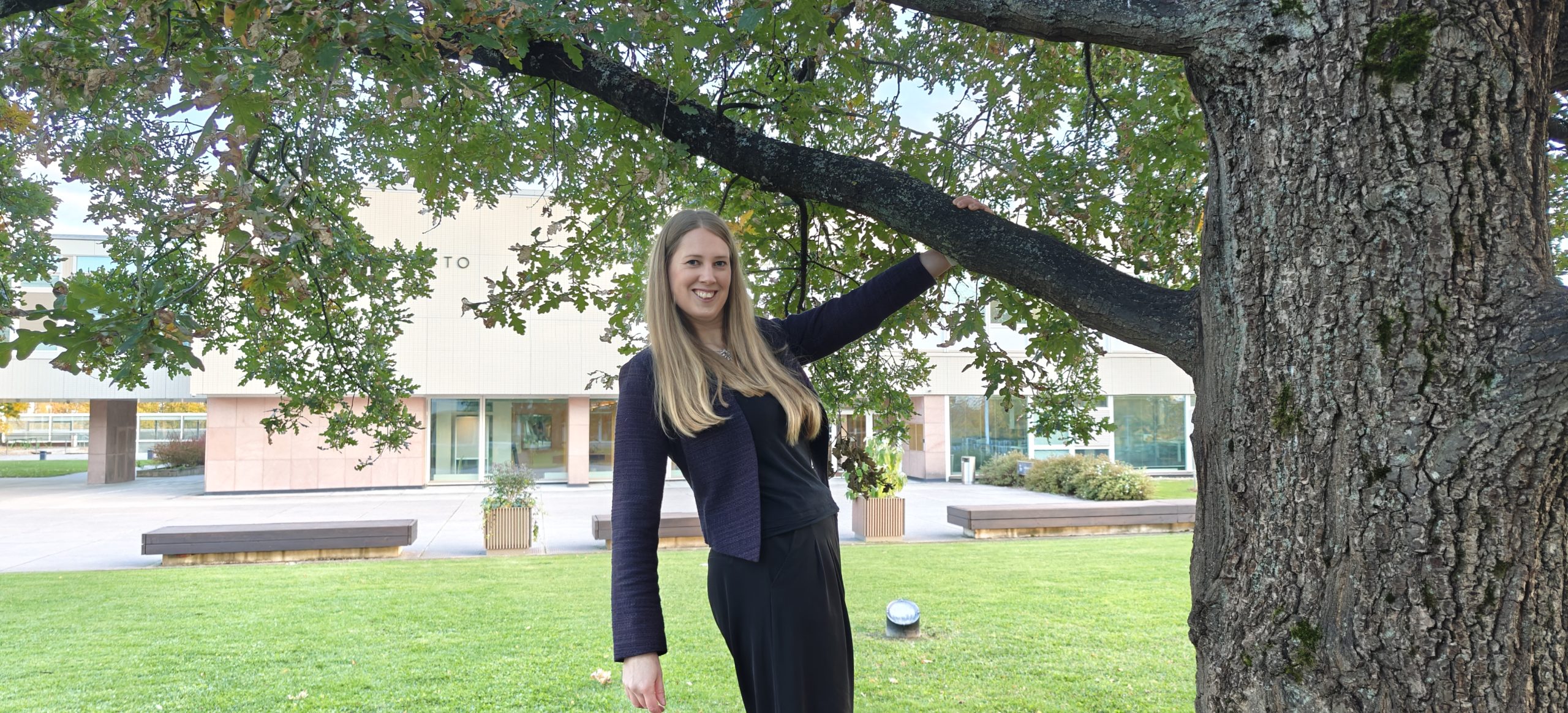 Elina yliopiston keskusaukiolla, päärakennus taustalla, käsi puun oksasta kiinni